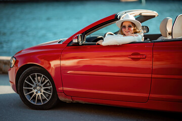 Outdoor summer portrait of stylish blonde woman driving red car convertible. Fashionable attractive woman with blond hair in a white hat in a red car. Sunny bright colors taken outdoors against the