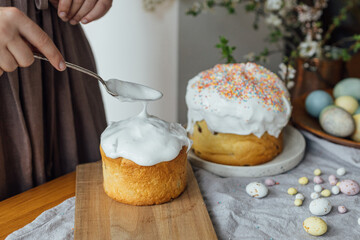 Woman decorating homemade easter bread with sugar glaze on rustic table with natural dyed eggs and spring blossom in room. Happy Easter! Woman baking stylish easter cake