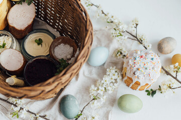 Obraz na płótnie Canvas Stylish easter bread and natural dyed eggs on rustic table with spring blossom and wicker basket. Traditional Easter basket food - easter cake with icing and sprinkles, eggs, ham, beets, butter.