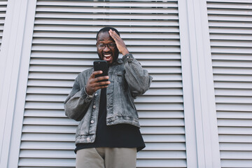 Excited male in optical spectacles surprised with good news in text message using cellphone technology in city, amazed hipster guy received coupon discount for making travel booking via mobile app