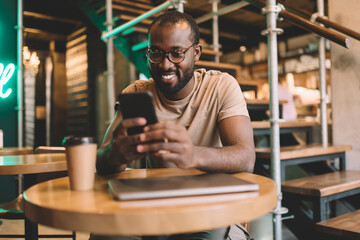 Happy African American male blogger in eyewear smiling while creating content plan in coworking space, cheerful hipster guy with modern cellular technology enjoying web chatting in social media