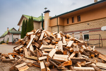 A pile of split firewood for heating the house, unloaded in the yard, against the backdrop of the house, natural heating sources.