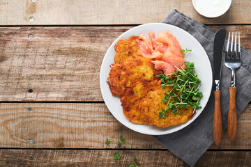 Potato pancakes. Fried homemade potato pancakes or latkes with cream, green onions, microgreens, red salmon and sauce in rustic plate on old wooden table background. Rustic style. Top view.