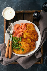 Potato pancakes. Fried homemade potato pancakes or latkes with cream, green onions, microgreens, red salmon and sauce in rustic plate on old black table background. Rustic style. Top view.