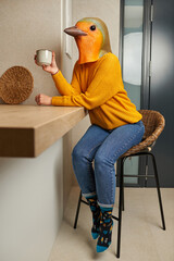 A woman dressed in a bird mask drinks a cup of coffee in the kitchen