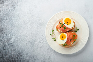Sandwich with delicious toppings on top: smoked salmon, eggs, herbs and microgreens radish, black sesame seeds over white plate on concrete background. Healthy open sandwich superfood. Top view.