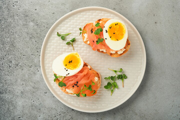 Sandwich with delicious toppings smoked salmon, eggs, herbs and microgreens radish, black sesame seeds over white plate on gray concrete table background. Healthy open sandwich superfood. Top view.