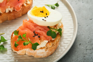 Sandwich with delicious toppings smoked salmon, eggs, herbs and microgreens radish, black sesame seeds over white plate on gray concrete table background. Healthy open sandwich superfood. Top view.