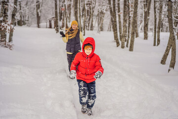 Fototapeta na wymiar Funny little boy and his mom in blue winter clothes walks during a snowfall. Outdoors winter activities for kids. Cute child wearing a warm hat low over his eyes catching snowflakes with his tongue
