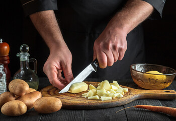 Cook cuts raw potatoes into pieces with a knife before preparing breakfast or dinner. Close-up of chef hands while working in kitchen