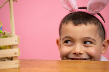 Joyful little boy with bunny ears on head looking from under the table with sly eyes at the basket...