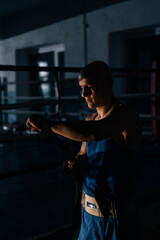 Vertical shot of confident boxer wrapping defense boxing tape around arms before fight in sport club with dark interior on background of ring. Serious fighter getting ready for fight in fitness center