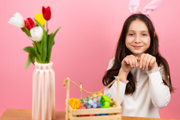 Pretty little girl wearing bunny ears on head folded her hands like rabbit sitting at the table with basket and vase with flowers and looking at the camera.