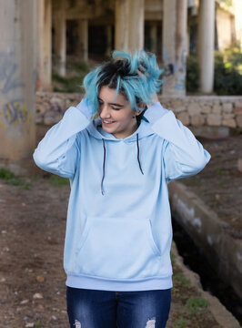 Blue haired Smiling Teenage girl in light blue oversize hoodie stays under bridge