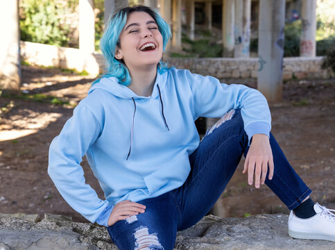 Laughing Blue Haired Teenage Girl In Light Blue Hoodie Sitting Under Bridge