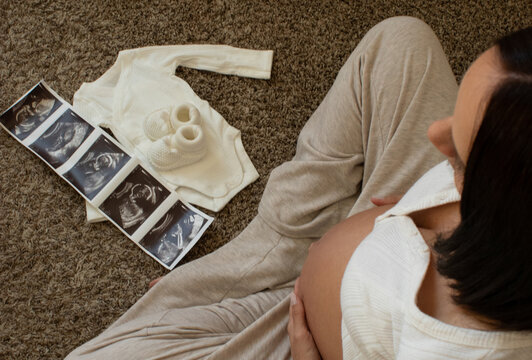 upper view of young woman sitting on the floor with ultrasound pictures and baby cloths, holding belly