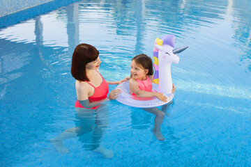 Little girl with unicorn inflatable ring with her mom in the same pink swimsuits relaxing in a swiming pool at sunny day. Happy mother and daughter in the pool at the leisure center.