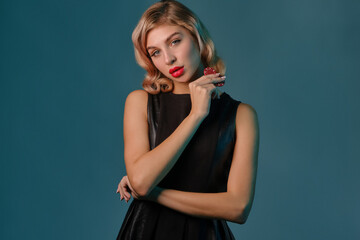 Naklejka premium Blonde girl in black leather dress showing two red chips, posing against blue background. Gambling entertainment, poker, casino. Close-up.