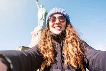 Young woman taking a selfie smiling with the statue of liberty in the background while sightseeing....
