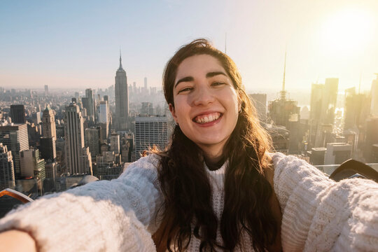 Young woman taking a selfie smiling on a rooftop with the empire state building in the background during sunset. Travel concept. influencer concept. Happiness concept.