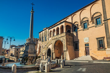 Tarquinia, Viterbo, Lazio, Italy - The main square of the village. The circular fountain with the obelisk and the cross. The town hall illuminated by the sun, in the blue sky. - obrazy, fototapety, plakaty