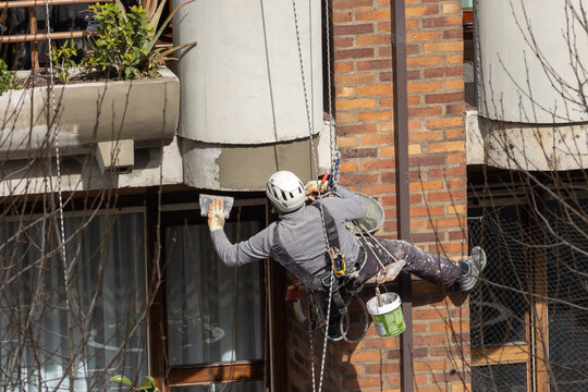 trabajador en altura realizando un trabajo vertical de mantenimiento de fachada estando colgado de cuerdas y llevando un casco y un arn&eacute;s de seguridad