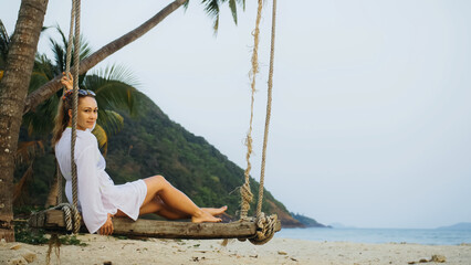 Fototapeta na wymiar Sensual barefoot woman on the swing on tropical beach. The girl in white shirt enjoy life and has fun. Paradise beaches, white sand, palm trees. Evening warm sunset. Summer holiday vacation concept
