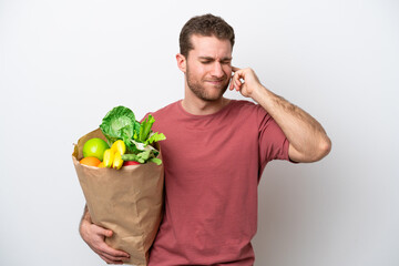 Young caucasian man holding a grocery shopping bag isolated on white background frustrated and covering ears