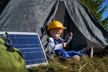 Close up of young boy with yellow helmet sitting in tent and watching at phone. Solar panel nearby...