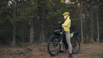 Fototapeta na wymiar The woman travel on mixed terrain cycle touring with bike bikepacking outdoor. The traveler journey with bicycle bags. Sportswear in green black colors. Magic forest park. Make a selfie smartphone.