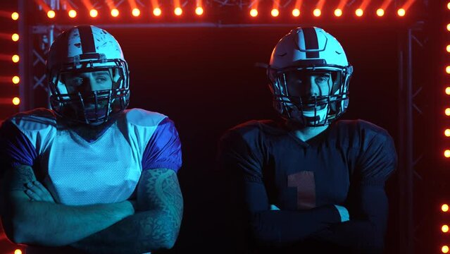 Two confident professional quarterbacks stand on the field against a backdrop of red light and smoke. American football players before the game match. Close up. Slow motion.