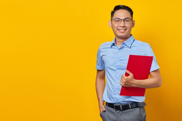 Portrait of handsome smiling young man in glasses holding document folder isolated on yellow...