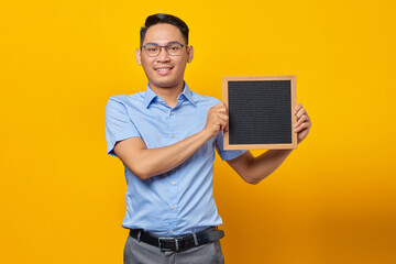 Portrait of smiling young Asian businessman in glasses holding chalkboard and standing confident looking at camera isolated over yellow background