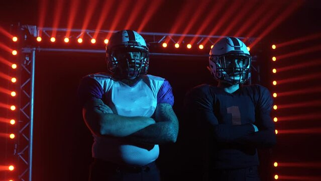 Two confident professional quarterbacks stand on the field against a backdrop of red light and smoke. American football players before the game match. Close up. Slow motion.
