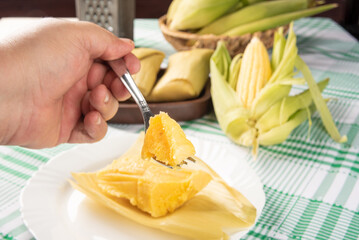 Hand holding a piece of Brazilian corn snack pamonha and green corn arranged on a table with green and white tablecloth, selective focus.