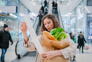 Rising food prices. Surprised woman looking into a paper check at the mall, holding a paper bag with fresh herbs and a baguette. - obrazy, fototapety, plakaty