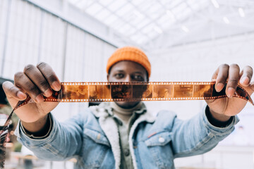 Emotional happy African-American man photographer in denim jacket looks at vintage film in brightly lit room extreme closeup - obrazy, fototapety, plakaty