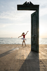 Female ballet dancer is posing near art object on seafront during sunrise