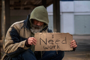 Poorly dressed gray-haired homeless beggar man holding a piece of cardboard with the inscription I...