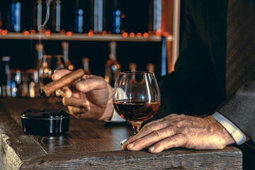 Fototapeta na wymiar Man's hands with a cigar, elegant glass of brandy on the bar counter. Alcoholic drinks, cognac, whiskey, port, brandy, rum, scotch, bourbon. Vintage wooden table in a pub at night