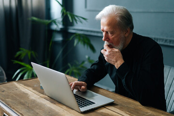 Close-up of thoughtful worried senior aged businessman working on laptop at workplace in dark home office room. Side view of concentrated mature adult male using computer looking on screen. - obrazy, fototapety, plakaty