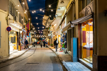 A colorfully illuminated narrow street of shops and cafes in the busy and touristic Plaka district of Athens, Greece at night.