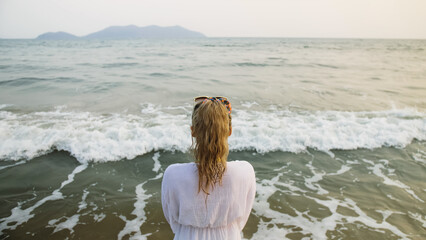 Blur mist glow cinematic portrait woman in a white tunic shirt on beach, near stormy sea. Attractive girl with wet curly hair, in sunglasses. Female tourist walks has fun rest, rear view