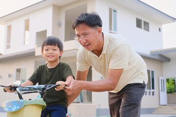Asian Father and son having fun learning to ride a bicycle in front of their new home, Dad and son spend time together, Dad tech little kid to ride bike, father's day, cycling for children and family