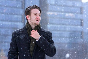 Portrait of handsome happy cheerful positive European blonde guy, young man office worker in falling snow is going to his job, work and smiling at cold winter snowy day in scarf, coat. 