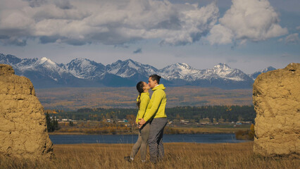 Man and woman in yellow green sportswear. Lovely couple of travelers hug and kiss near old stone enjoying highland landscape. Two travelers are walking against the backdrop of snow-capped mountains.