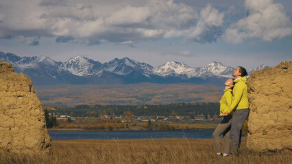 Man and woman in yellow green sportswear. Lovely couple of travelers hug and kiss near old stone enjoying highland landscape. Two travelers are walking against the backdrop of snow-capped mountains.