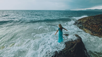 Sexy hot woman meditates, relaxes on a rock reef hill in stormy morning rain cloudy sea. Girl in blue swimsuit, dress tunic. Concept feminine, relax, sexual health. Dark dramatic silhouette view
