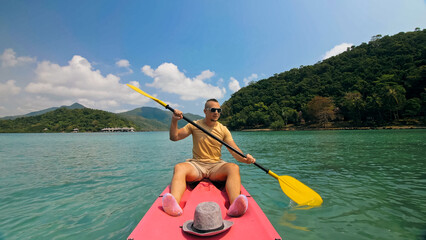Young man with sunglasses and hat rows pink plastic canoe along sea against green hilly islands...