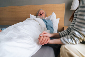 Close-up of unrecognizable loving young woman child holding hand of sick aged father lying in hospital bed. Aged grandfather male patient resting at home and holding hand of granddaughter visitor.